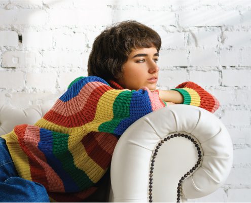 April Blog 2021 young woman leaning on a white couch wearing a rainbow knitted sweater